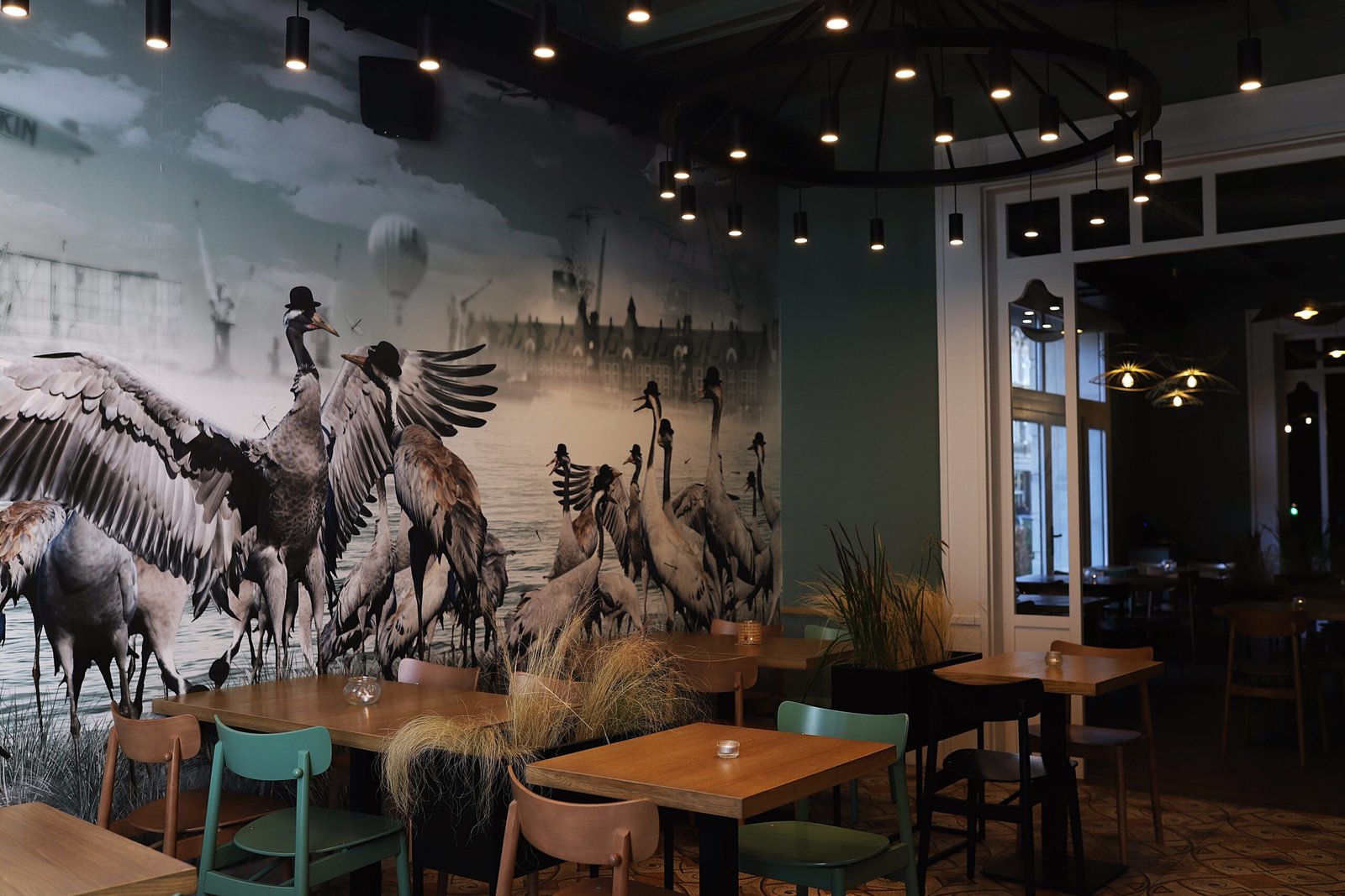 Mural z żurawiami na ścianie, lampy wiszące i dębowe stoły z miętowymi krzesłami