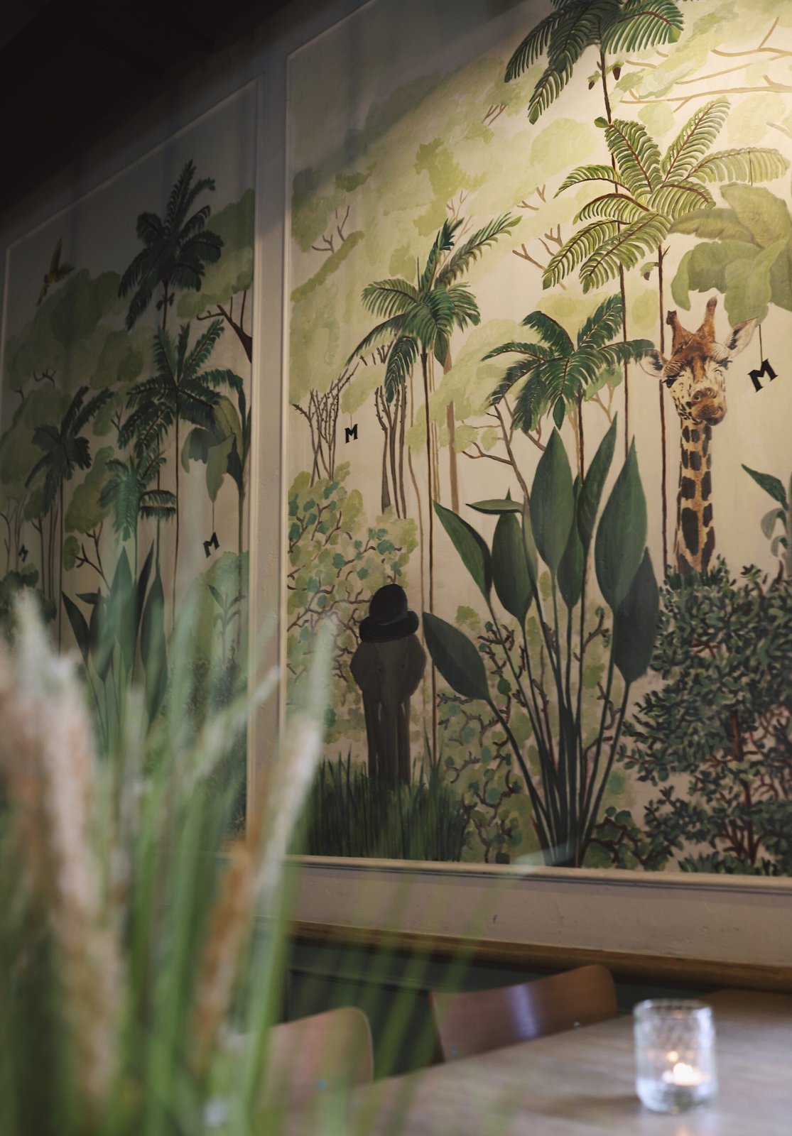 Ręcznie malowany mural dżungli z żyrafą, palmami i małą literą M ukrytą w liściach